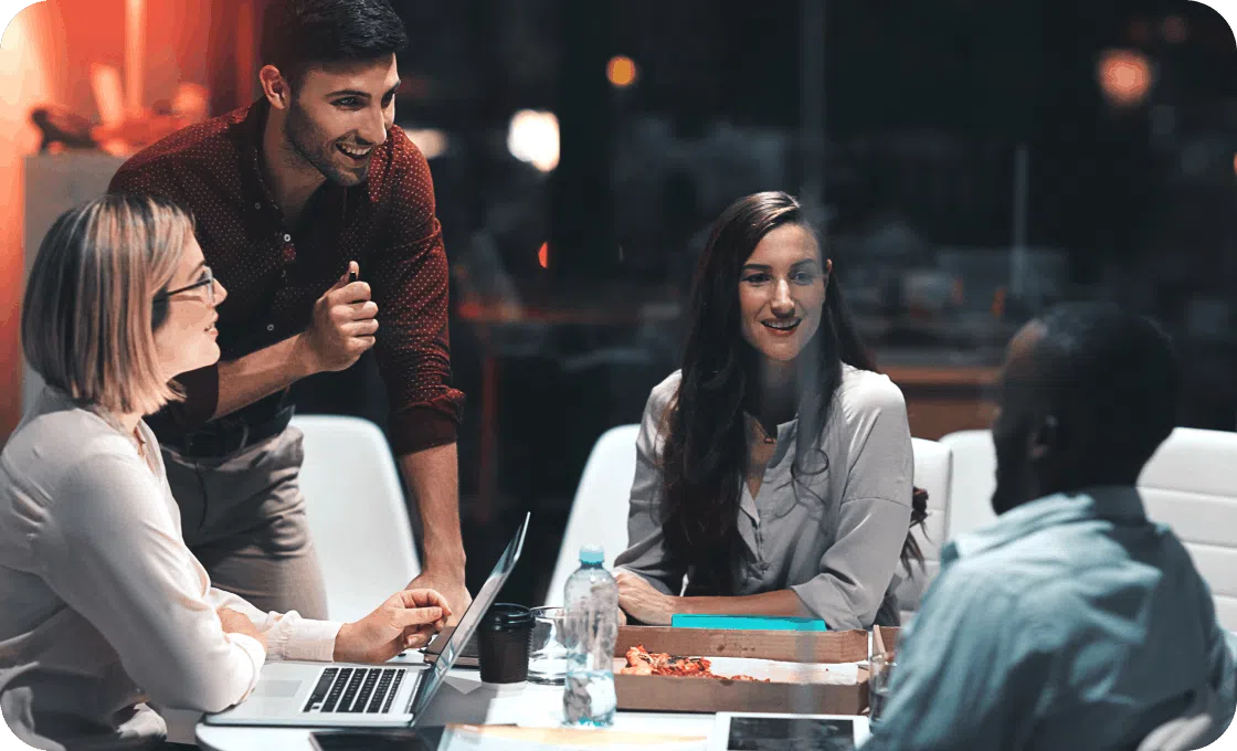 A diverse group of four colleagues is engaged in a lively discussion around a table with laptops and papers. The setting is an office, evoking a collaborative and positive atmosphere.