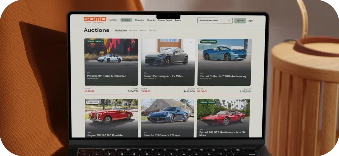 A laptop displays an online car auction page with listings of various luxury cars. The background has warm brown tones, creating a cozy atmosphere.