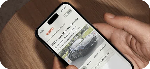 A person holding a smartphone displays a website with a 2023 Porsche 911 Turbo S Cabriolet. The screen shows auction details and a car image.