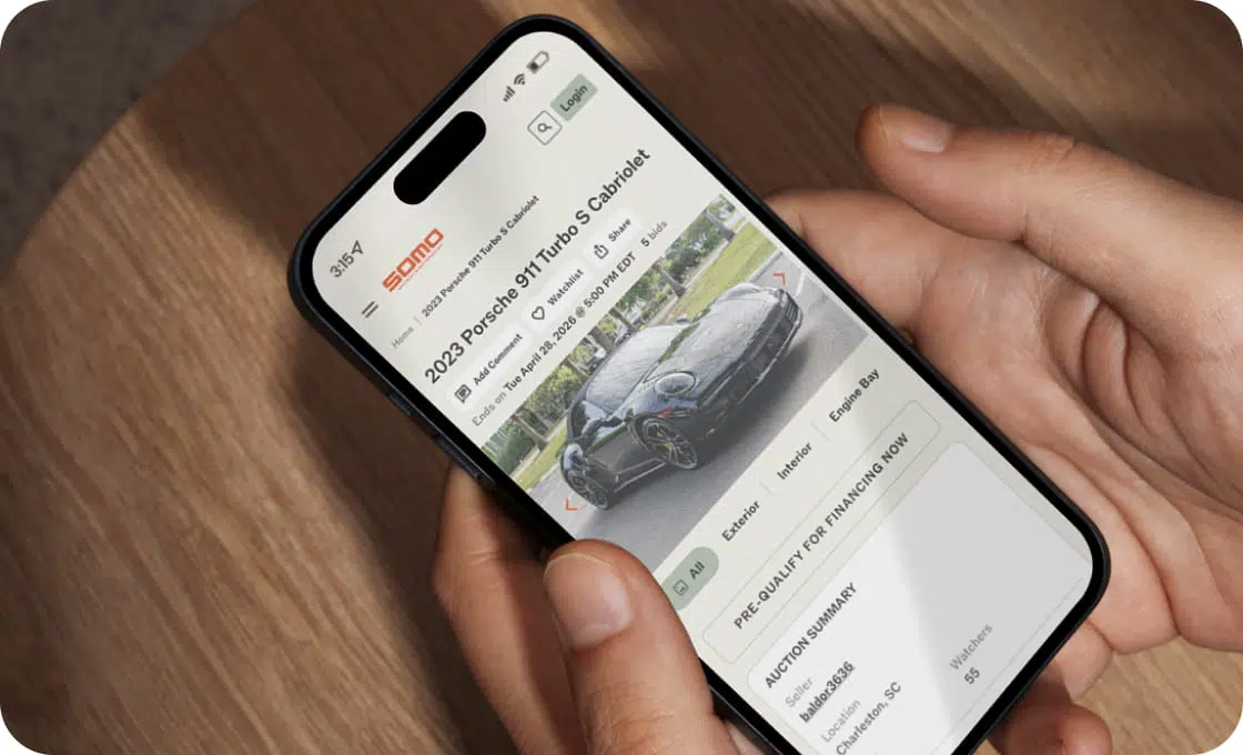 A person holding a smartphone displays a website with a 2023 Porsche 911 Turbo S Cabriolet. The screen shows auction details and a car image.